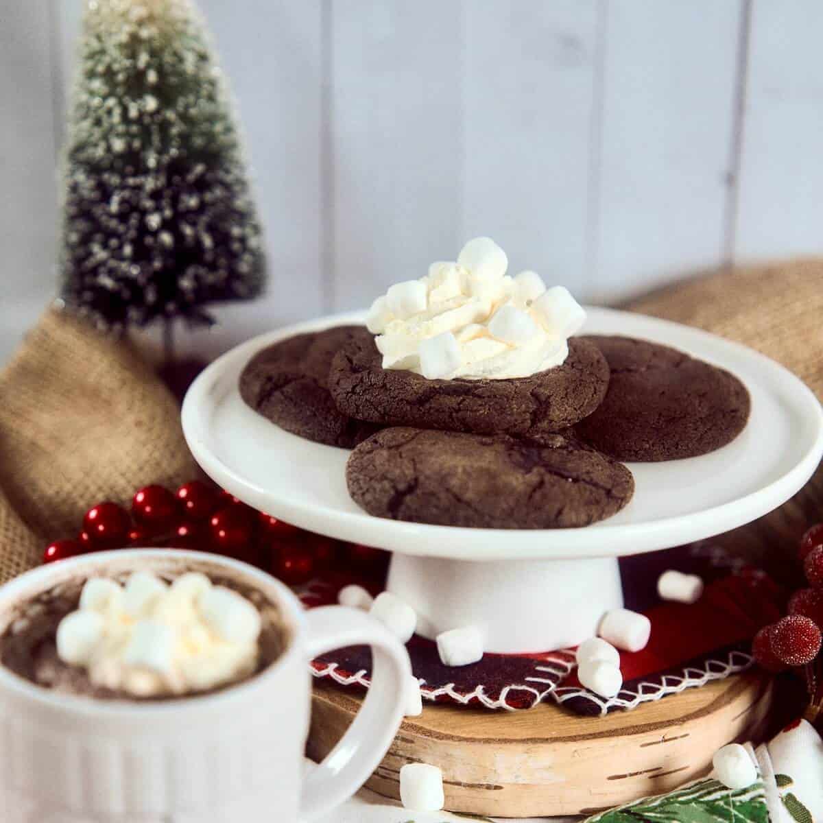 Hot Chocolate Lava Cookies stacked on a white cake stand with a cup hot chocolate in front.