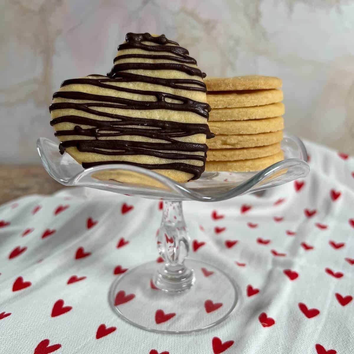 Heart shaped shortbread cutout cookies stacked on a small cake stand with one standing up drizzled with chocolate.