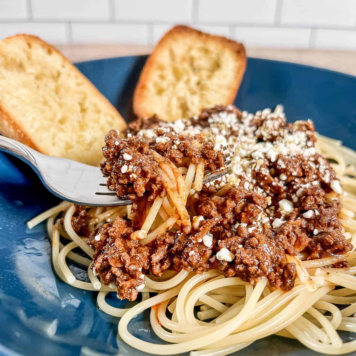 Fork lifting a portion of spaghetti topped with Cincinnati chili and feta cheese with two pieces of garlic bread.