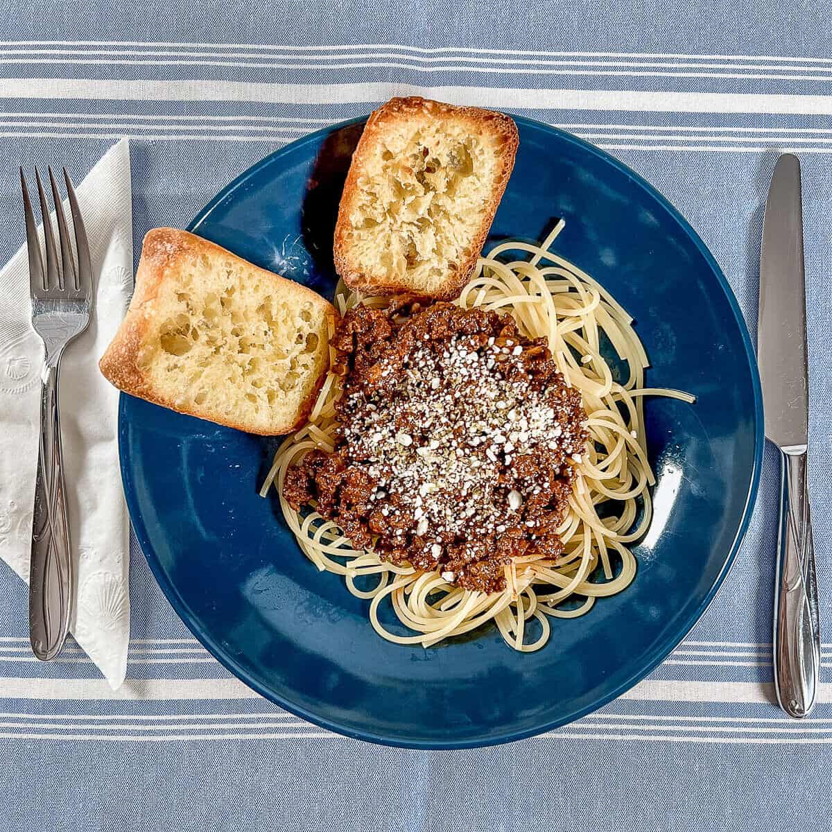 Cincinnati chili on a bowl of spaghetti topped with feta cheese with two pieces of garlic bread surrounded by a fork and knife.