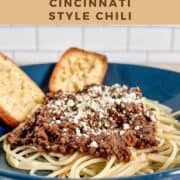 Cincinnati chili on a bowl of spaghetti topped with feta cheese with two pieces of garlic bread Pinterest banner.