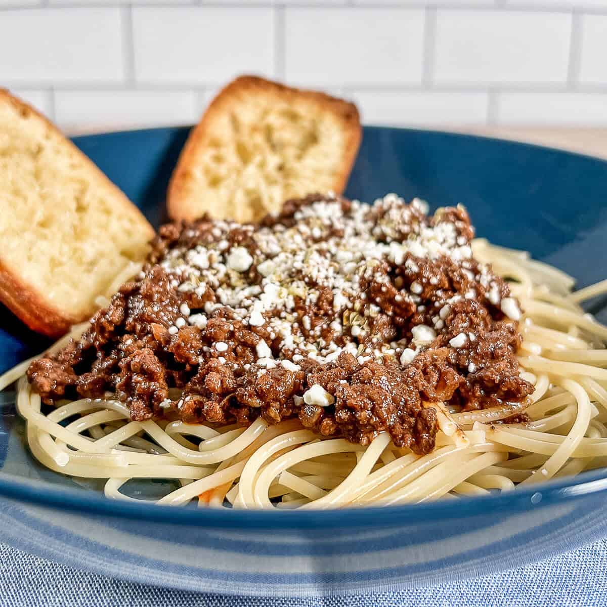Cincinnati chili on a bowl of spaghetti topped with feta cheese with two pieces of garlic bread.