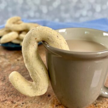 A cup of tea with a almond shortbread cookie hanging on the mug with a plateful behind.