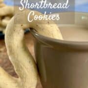 A cup of tea with a almond shortbread cookie hanging on the mug with a plateful behind Pinterest banner.