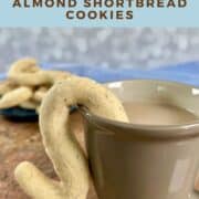 A cup of tea with a almond shortbread cookie hanging on the mug with a plateful behind Pinterest banner.