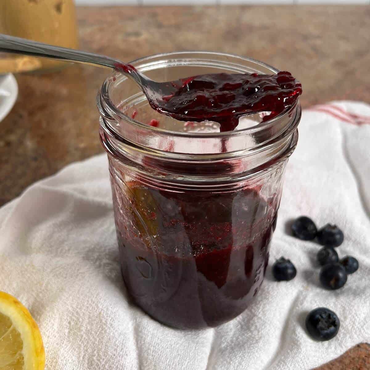A spoonful of jam above a jar of jam next to lemons and blueberries.