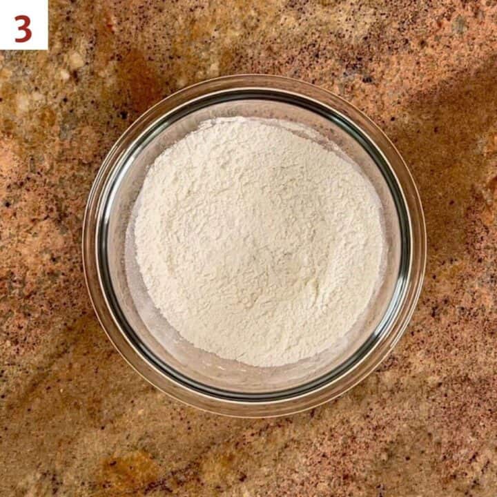 A bowl of flour, baking power, and salt on a marble counter.
