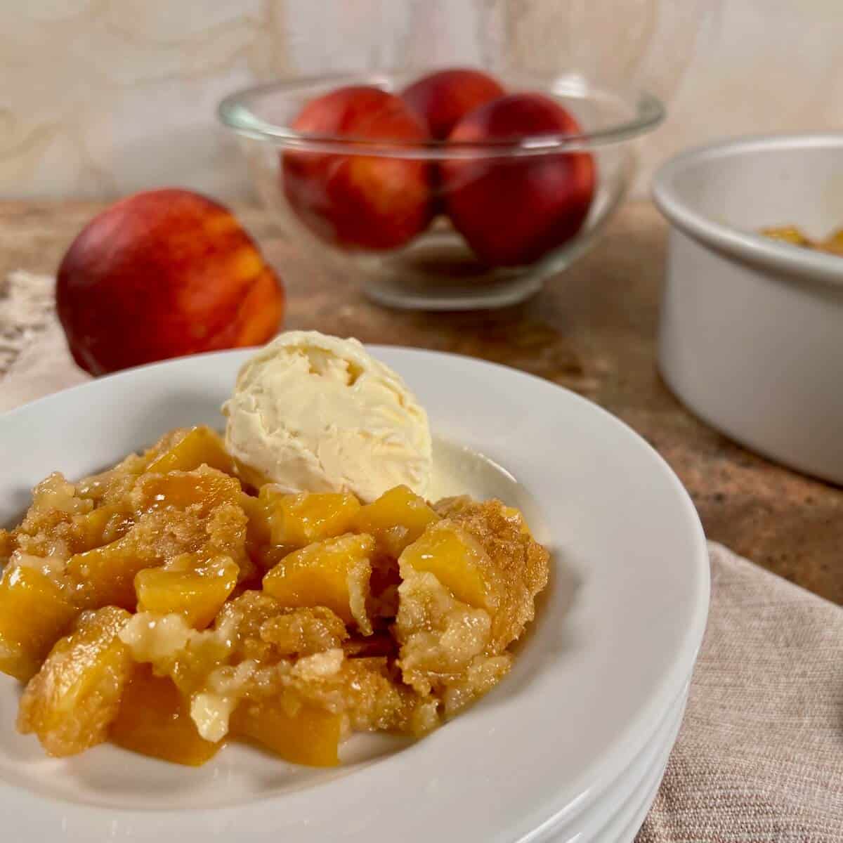 Peach cobbler with vanilla ice cream on stacked white plates with peaches behind.