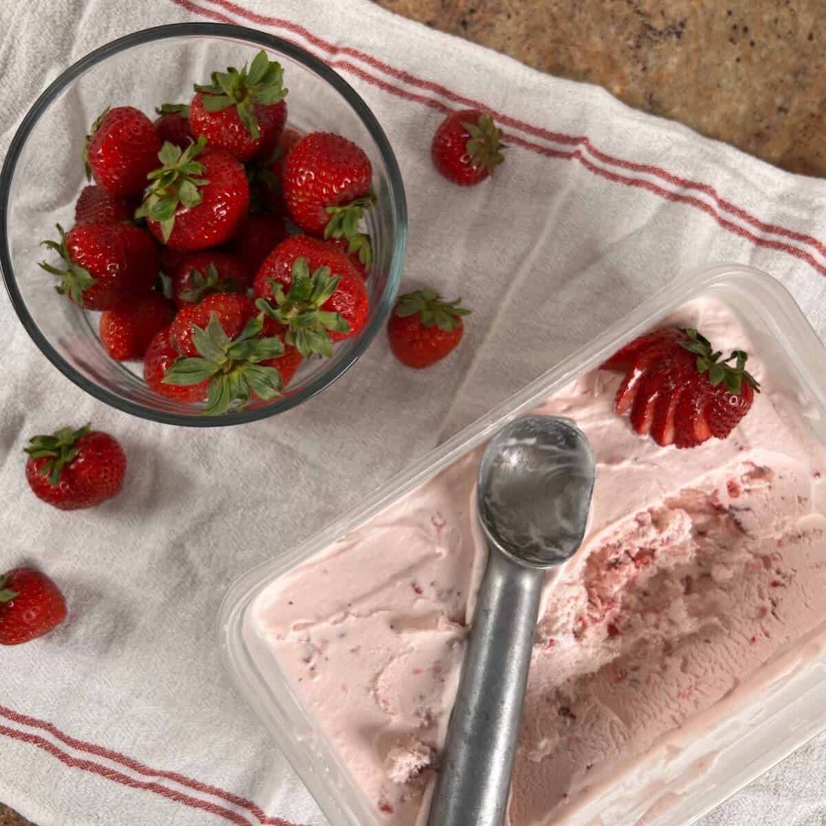 An ice cream scoop in a container of strawberry ice cream next to a glass bowl of strawberries.