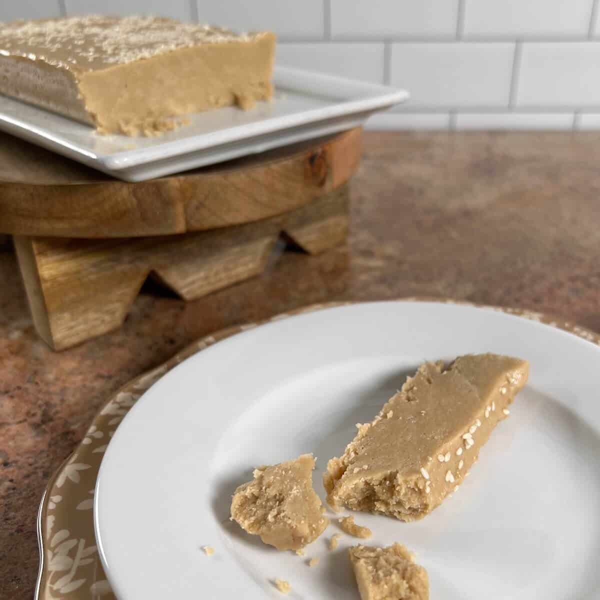 A piece of Tahini Halva on a white plate with the slab on a raised tray behind.