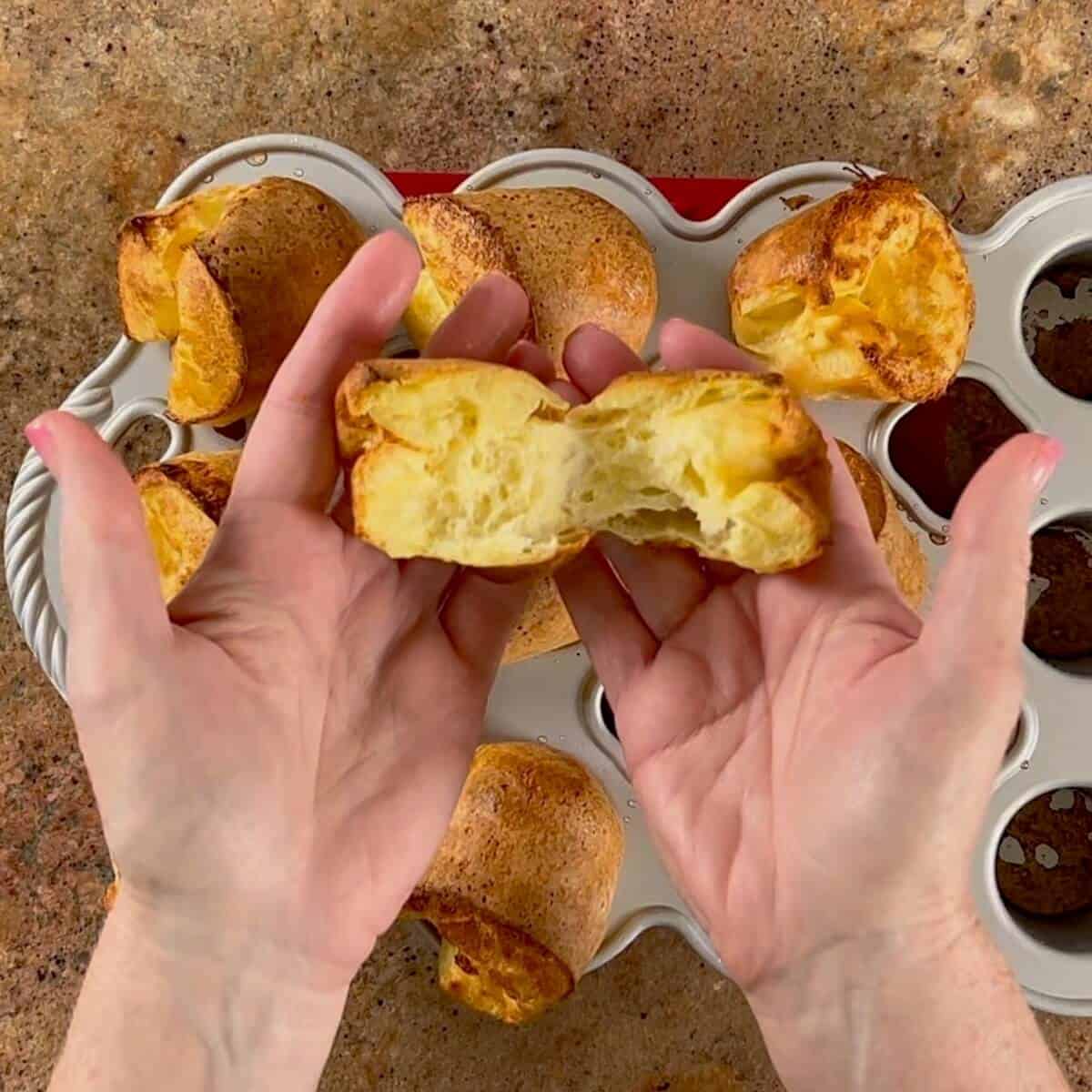 Fluffy golden popover with buttery interior in hands over a pan of baked popovers.