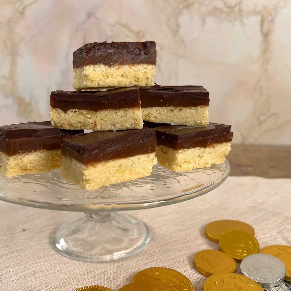 A stacked pile of Millionaire's Shortbread on a glass cake stand with chocolate coins below.