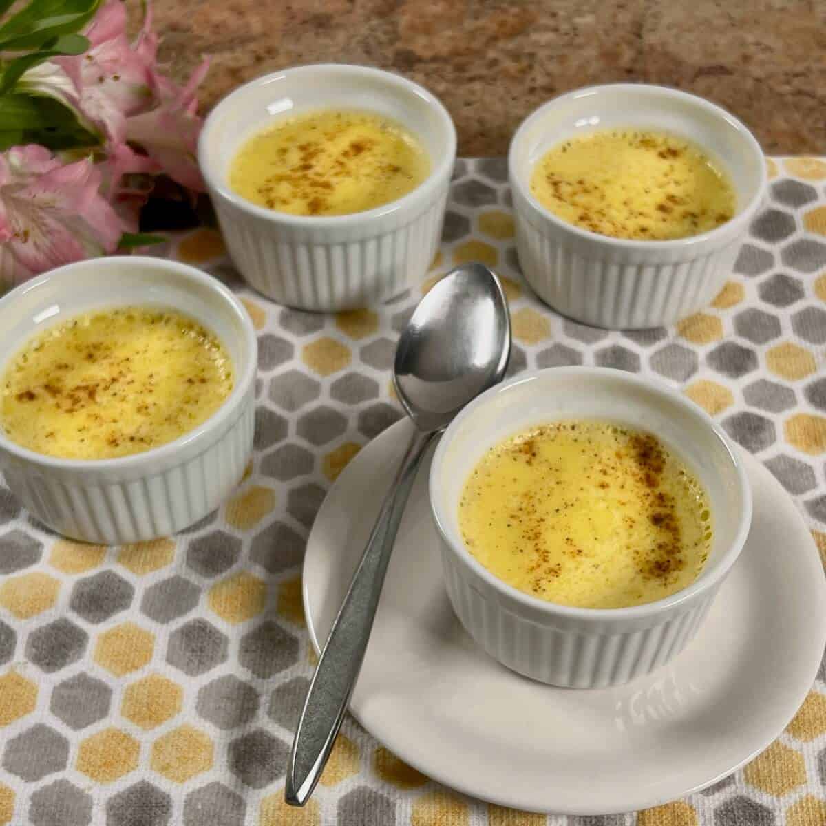 Four ramekins of egg custard set on a patterned cloth with a spoon and pink flowers nearby.