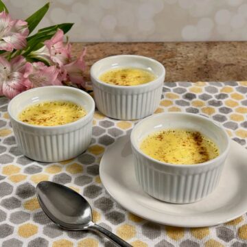 Three ramekins of egg custard set on a patterned cloth with a spoon and pink flowers nearby.