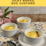 Three ramekins of egg custard set on a patterned cloth with a spoon and pink flowers nearby Pinterest banner.