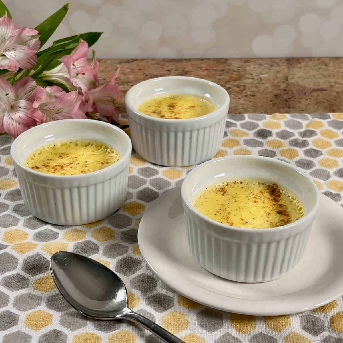 Three ramekins of egg custard set on a patterned cloth with a spoon and pink flowers nearby.