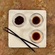 Vanilla bean powder, vanilla bean paste, vanilla extract, and 2 whole vanilla beans on a white plate.