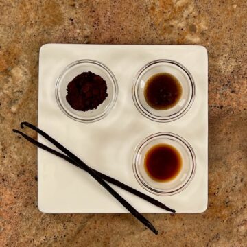 Vanilla bean powder, vanilla bean paste, vanilla extract, and 2 whole vanilla beans on a white plate.